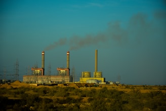 An industrial facility with multiple smokestacks emitting smoke into a clear blue sky, surrounded by power lines and a barren, desert-like landscape in the foreground.