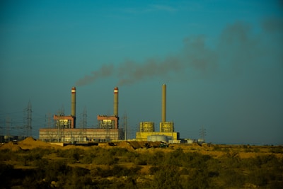 An industrial facility with multiple smokestacks emitting smoke into a clear blue sky, surrounded by power lines and a barren, desert-like landscape in the foreground.