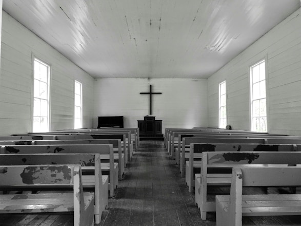 A simple, wooden church interior with rows of empty, worn pews facing a central altar. A large cross hangs on the wall above a minimalist pulpit against a backdrop of muted, aged wood. Natural light softly filters through tall, rectangular windows lining both sides of the space, creating a serene and contemplative atmosphere.