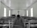 A simple, wooden church interior with rows of empty, worn pews facing a central altar. A large cross hangs on the wall above a minimalist pulpit against a backdrop of muted, aged wood. Natural light softly filters through tall, rectangular windows lining both sides of the space, creating a serene and contemplative atmosphere.