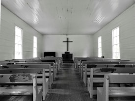 A simple, wooden church interior with rows of empty, worn pews facing a central altar. A large cross hangs on the wall above a minimalist pulpit against a backdrop of muted, aged wood. Natural light softly filters through tall, rectangular windows lining both sides of the space, creating a serene and contemplative atmosphere.