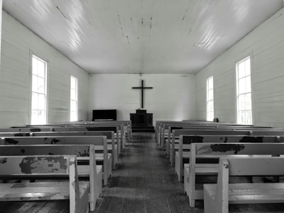 A simple, wooden church interior with rows of empty, worn pews facing a central altar. A large cross hangs on the wall above a minimalist pulpit against a backdrop of muted, aged wood. Natural light softly filters through tall, rectangular windows lining both sides of the space, creating a serene and contemplative atmosphere.