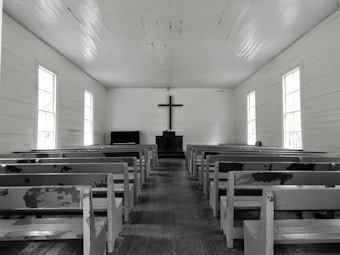 A simple, wooden church interior with rows of empty, worn pews facing a central altar. A large cross hangs on the wall above a minimalist pulpit against a backdrop of muted, aged wood. Natural light softly filters through tall, rectangular windows lining both sides of the space, creating a serene and contemplative atmosphere.