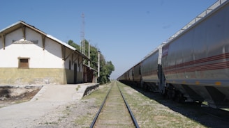 A railway scene featuring a long line of cargo train cars on tracks extending into the distance. On the left, there is a small, rustic building with a sign that reads 'C. Cienegas'. The ground is a mix of gravel and grass, and the sky is clear and blue.
