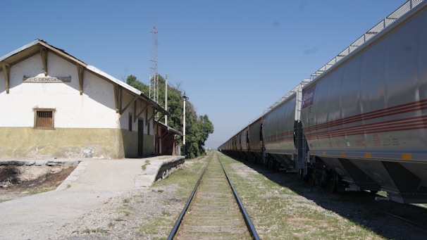 A railway scene featuring a long line of cargo train cars on tracks extending into the distance. On the left, there is a small, rustic building with a sign that reads 'C. Cienegas'. The ground is a mix of gravel and grass, and the sky is clear and blue.
