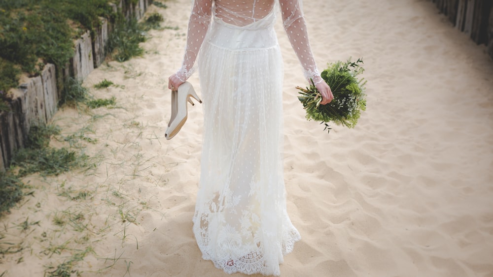 woman wearing white wedding dress walking on white sand holding pair of beige shoes and bouquet of flowers during day