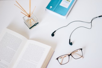 An open book lies on a white surface alongside a pair of glasses, a set of earphones, an aroma diffuser with reeds, and a closed blue notebook. The items are arranged in a casual manner, evoking a sense of study or relaxation.