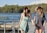 Two women wearing sundresses and wide-brimmed hats hold hands while walking on a wooden pier. They are smiling and appear to be enjoying a sunny day by a lake, with a backdrop of trees and a clear blue sky.