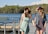 Two women wearing sundresses and wide-brimmed hats hold hands while walking on a wooden pier. They are smiling and appear to be enjoying a sunny day by a lake, with a backdrop of trees and a clear blue sky.