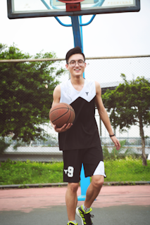 A person with glasses, wearing a black and white sports outfit is playing basketball on an outdoor court. He holds a basketball with one hand while smiling and walking towards the camera. The background includes a basketball hoop and greenery.