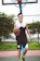 A person with glasses, wearing a black and white sports outfit is playing basketball on an outdoor court. He holds a basketball with one hand while smiling and walking towards the camera. The background includes a basketball hoop and greenery.