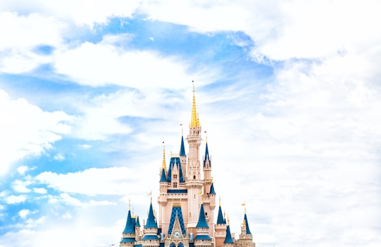 A majestic fairy tale castle with blue spires and golden accents stands against a bright, partly cloudy sky. The architecture is whimsical with intricate details typical of a fantasy theme park.
