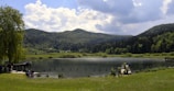 A serene lake surrounded by lush green hills and dense forests under a partly cloudy sky. People are gathered in small groups on the grassy banks and on wooden platforms built over the water.