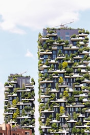 Two modern skyscrapers covered with abundant greenery, featuring plants and trees on every balcony. The buildings are architecturally unique, integrating nature into an urban setting. A clear sky with some clouds is visible in the background.
