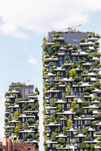 Two modern skyscrapers covered with abundant greenery, featuring plants and trees on every balcony. The buildings are architecturally unique, integrating nature into an urban setting. A clear sky with some clouds is visible in the background.