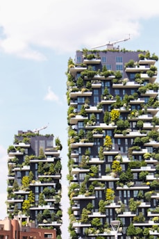 Two modern skyscrapers covered with abundant greenery, featuring plants and trees on every balcony. The buildings are architecturally unique, integrating nature into an urban setting. A clear sky with some clouds is visible in the background.