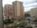 High-rise residential and commercial buildings dominate an urban landscape. The scene includes a mix of older and modern architecture with prominent balconies. A landscaped area with trees and bushes is visible, along with commercial establishments at street level. The sky is overcast, casting a neutral light over the scene.