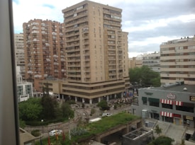 High-rise residential and commercial buildings dominate an urban landscape. The scene includes a mix of older and modern architecture with prominent balconies. A landscaped area with trees and bushes is visible, along with commercial establishments at street level. The sky is overcast, casting a neutral light over the scene.