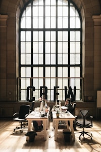 A spacious, well-lit office space with large arched windows letting in natural light. The room features several desks equipped with computers, office chairs, houseplants, and various office supplies. The word 'CREW' is prominently displayed in large black letters on the window sill. There are a few people working at the desks.
