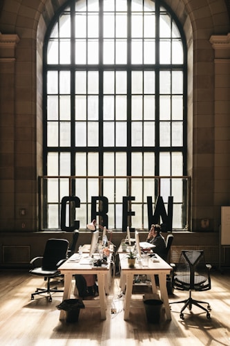 A spacious, well-lit office space with large arched windows letting in natural light. The room features several desks equipped with computers, office chairs, houseplants, and various office supplies. The word 'CREW' is prominently displayed in large black letters on the window sill. There are a few people working at the desks.