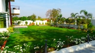 A neatly landscaped garden with lush green grass and various flowering plants surrounding the perimeter. Palm trees are spaced evenly along a white concrete wall, adding a tropical feel. In the foreground, a variety of small white flowers are in bloom, while a modern house with a glass facade is partially visible on the left. The sky is clear and blue, suggesting a sunny day.