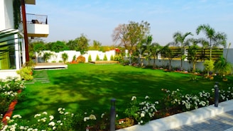 A neatly landscaped garden with lush green grass and various flowering plants surrounding the perimeter. Palm trees are spaced evenly along a white concrete wall, adding a tropical feel. In the foreground, a variety of small white flowers are in bloom, while a modern house with a glass facade is partially visible on the left. The sky is clear and blue, suggesting a sunny day.
