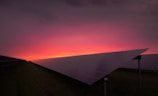 A field of solar panels is visible with a vibrant sunset in the background. The panels are arranged in rows, and the sky transitions from a warm pink and orange near the horizon to a dark purple at the top.