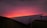 A field of solar panels is visible with a vibrant sunset in the background. The panels are arranged in rows, and the sky transitions from a warm pink and orange near the horizon to a dark purple at the top.