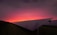 A field of solar panels is visible with a vibrant sunset in the background. The panels are arranged in rows, and the sky transitions from a warm pink and orange near the horizon to a dark purple at the top.