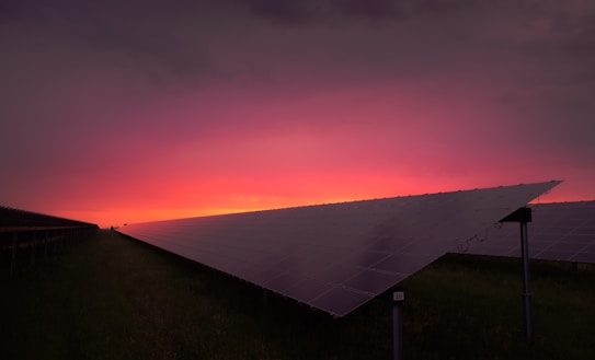 A field of solar panels is visible with a vibrant sunset in the background. The panels are arranged in rows, and the sky transitions from a warm pink and orange near the horizon to a dark purple at the top.