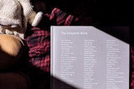 A book titled 'The Unsplash Book' lies on a surface alongside a pair of knitted gloves and a woolen red and black plaid scarf. A pair of tan leather boots is partially visible on the left. The book cover has a list of names printed in white text and is partially illuminated by sunlight, creating a shadow pattern.