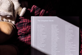A book titled 'The Unsplash Book' lies on a surface alongside a pair of knitted gloves and a woolen red and black plaid scarf. A pair of tan leather boots is partially visible on the left. The book cover has a list of names printed in white text and is partially illuminated by sunlight, creating a shadow pattern.