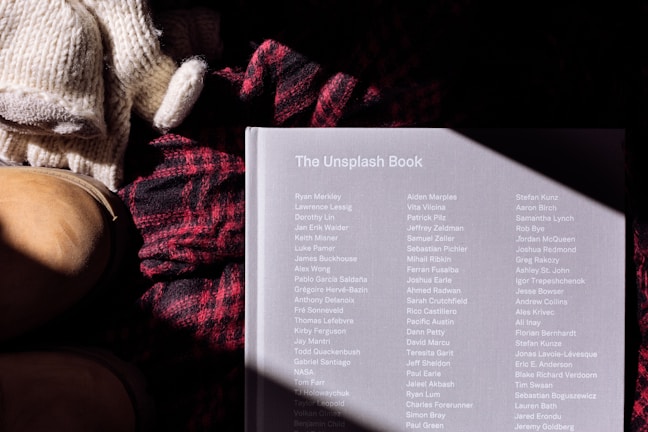 A book titled 'The Unsplash Book' lies on a surface alongside a pair of knitted gloves and a woolen red and black plaid scarf. A pair of tan leather boots is partially visible on the left. The book cover has a list of names printed in white text and is partially illuminated by sunlight, creating a shadow pattern.