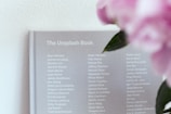A grey book titled 'The Unsplash Book' displays a list of names in white text on its cover. To the right, blurred pink flowers with green leaves are visible, adding a touch of color.