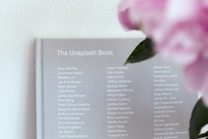 A grey book titled 'The Unsplash Book' displays a list of names in white text on its cover. To the right, blurred pink flowers with green leaves are visible, adding a touch of color.