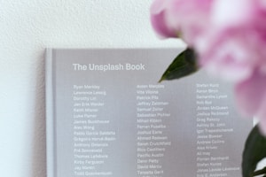 A grey book titled 'The Unsplash Book' displays a list of names in white text on its cover. To the right, blurred pink flowers with green leaves are visible, adding a touch of color.