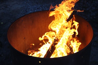 A metal barrel containing a bright, active fire with flames that are vibrant and orange, casting light on the immediate surroundings. The background is dimly lit, enhancing the warmth of the fire.