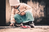 A young child wearing a knitted hat and sweater crouches down to play with a small red toy car on a rough, outdoor surface. The child is focusing intently while holding a small object, possibly a stone, in their hand. The background features a textured wall with a muted, rustic appearance.