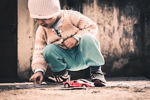 A young child wearing a knitted hat and sweater crouches down to play with a small red toy car on a rough, outdoor surface. The child is focusing intently while holding a small object, possibly a stone, in their hand. The background features a textured wall with a muted, rustic appearance.