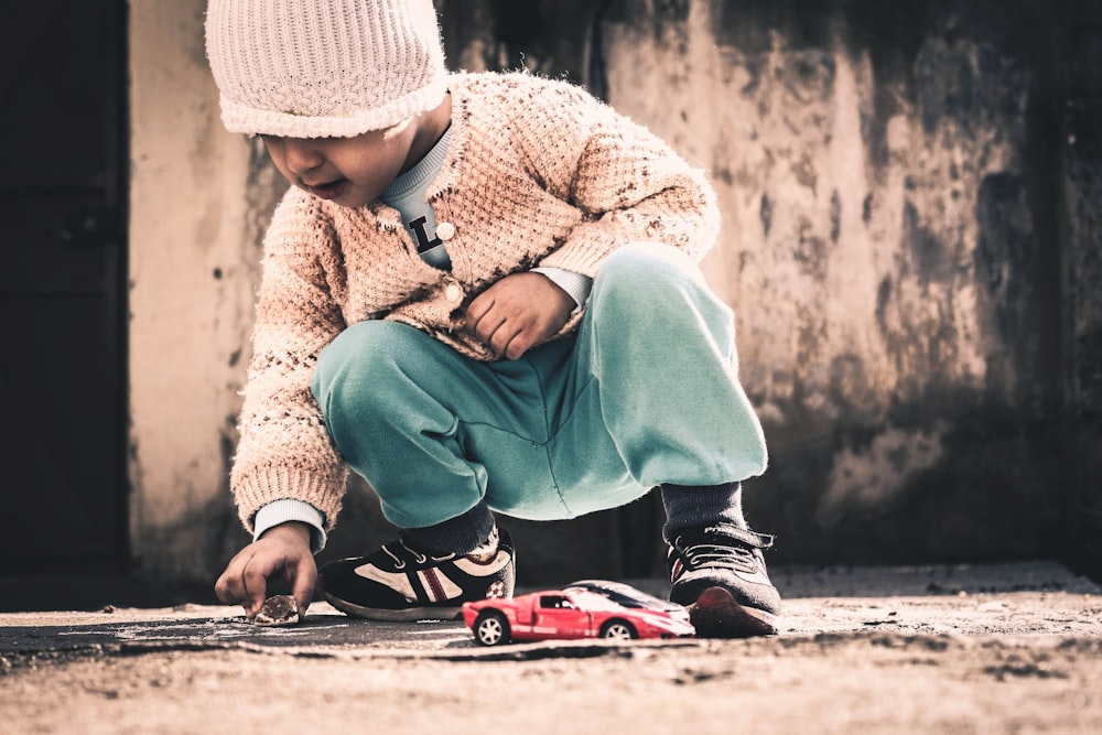 child squatting on beige floor holding rock near red sports coupe scale model at daytime