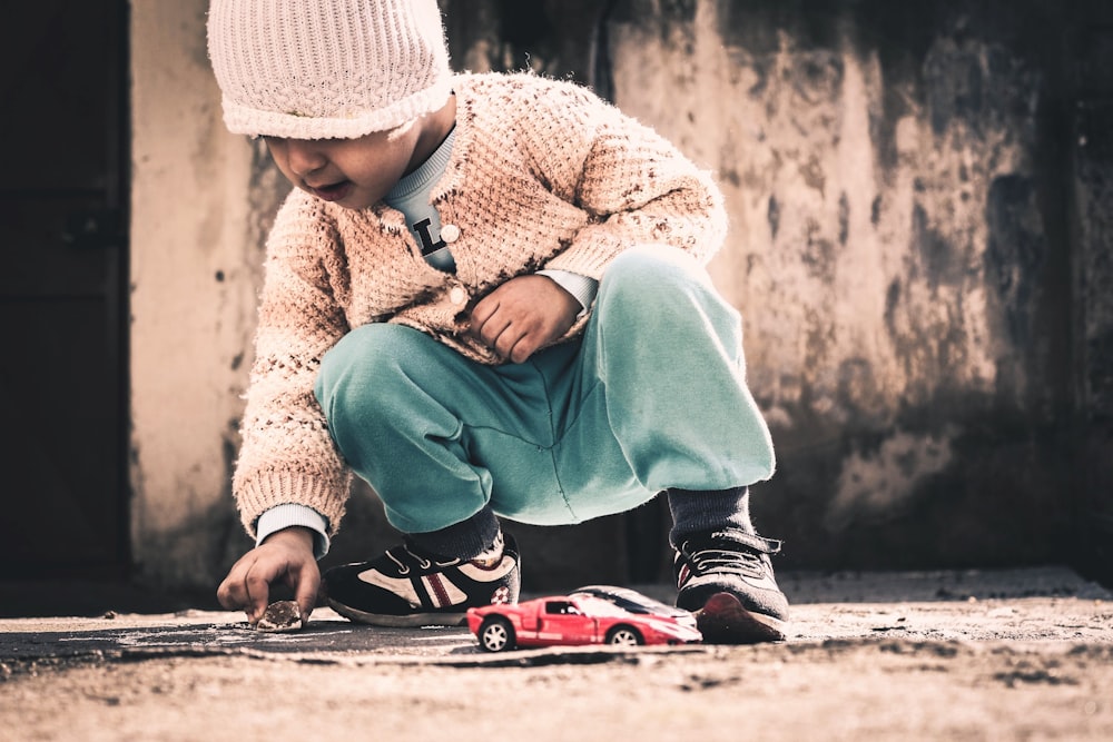 enfant accroupi sur le sol beige tenant un rocher près d’un coupé sport rouge modèle réduit à la journée