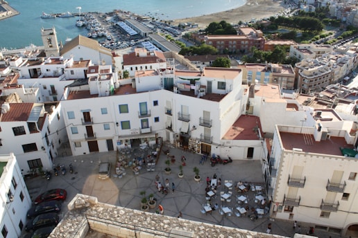 An aerial view of a coastal town featuring white buildings with red-tiled roofs and a courtyard filled with outdoor seating. The scene includes several parked cars, a marina with boats in the background, and the edge of a sandy beach. The area is bustling with people scattered throughout the courtyard, indicating a lively atmosphere.