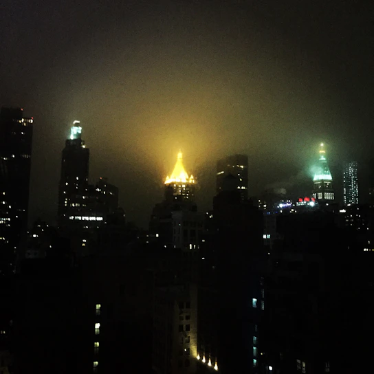 A moody, cinematic rooftop scene at night with soft gold lighting, an elegant crowd in silhouette, and a sparkling cityscape beyond.