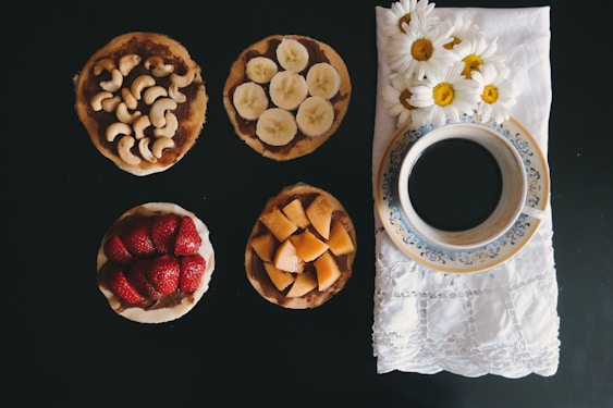 A selection of four small round snacks is arranged on a dark surface, each topped with different ingredients: cashews, banana slices, strawberries, and diced cantaloupe. To the right, a blue and white floral-patterned cup filled with black coffee sits on a white embroidered napkin adorned with fresh daisies.