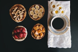 A selection of four small round snacks is arranged on a dark surface, each topped with different ingredients: cashews, banana slices, strawberries, and diced cantaloupe. To the right, a blue and white floral-patterned cup filled with black coffee sits on a white embroidered napkin adorned with fresh daisies.