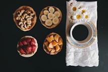 A selection of four small round snacks is arranged on a dark surface, each topped with different ingredients: cashews, banana slices, strawberries, and diced cantaloupe. To the right, a blue and white floral-patterned cup filled with black coffee sits on a white embroidered napkin adorned with fresh daisies.