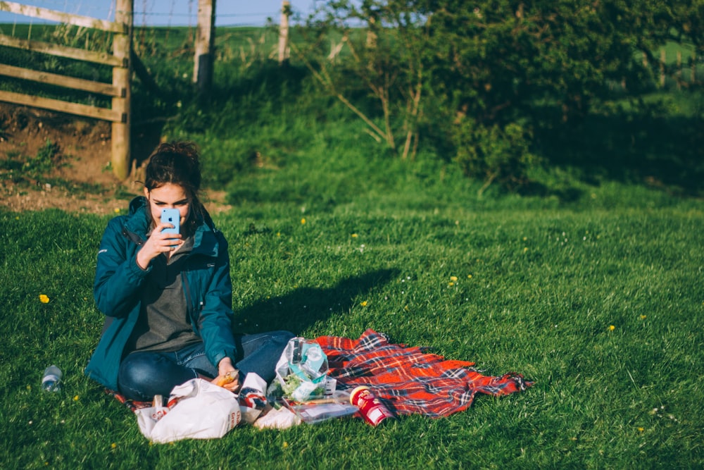 Mujer con atuendo azul usando un teléfono inteligente azul mientras está sentada en la hierba