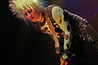 A person with spiky, bleach-blonde hair, wearing a dark eye makeup and a snake-patterned top. The style is punk-inspired, featuring a mesh sleeve with large holes and a belt adorned with chains.