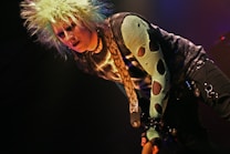 A person with spiky, bleach-blonde hair, wearing a dark eye makeup and a snake-patterned top. The style is punk-inspired, featuring a mesh sleeve with large holes and a belt adorned with chains.
