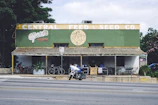 A vintage-style building with a sign reading 'Central Feed & Seed Co.' The structure has a green and yellow facade with a rustic, corrugated metal roof. Below, there is a taco bar named 'Güero's,' with outdoor seating where a few people are dining. A couple of bicycles are parked outside. A motorcycle is parked on the road in front of the building. Lush greenery surrounds the area, and there is an ATM machine visible on the right.